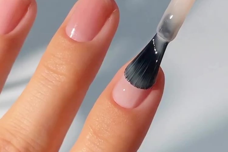 A imagem mostra uma mão com unhas pintadas em um tom rosa claro. Um pincel está aplicando uma camada de esmalte transparente sobre uma das unhas. A cena é iluminada de forma suave, destacando o brilho e a textura do esmalte. A aparência geral é de um cuidado delicado e bem executado nas unhas.