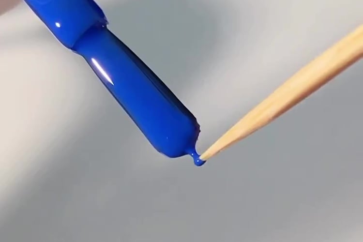 A imagem mostra um pincel de esmalte azul prestes a tocar a ponta de um palito de madeira. Uma gota de esmalte está se formando na extremidade do pincel, quase se conectando ao palito. O fundo é desfocado, destacando o contraste entre o azul vibrante do esmalte e o tom natural do palito.