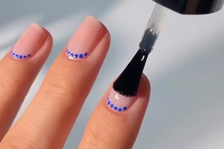 A imagem mostra uma mão com unhas decoradas de maneira delicada. As unhas têm um esmalte base claro, e cada uma delas possui uma linha de pequenas bolinhas azuis perto da ponta. Um pincel de esmalte está aplicando uma camada de acabamento transparente sobre o design, destacando o brilho e a nitidez das bolinhas. O fundo é suave, mantendo o foco nas unhas.