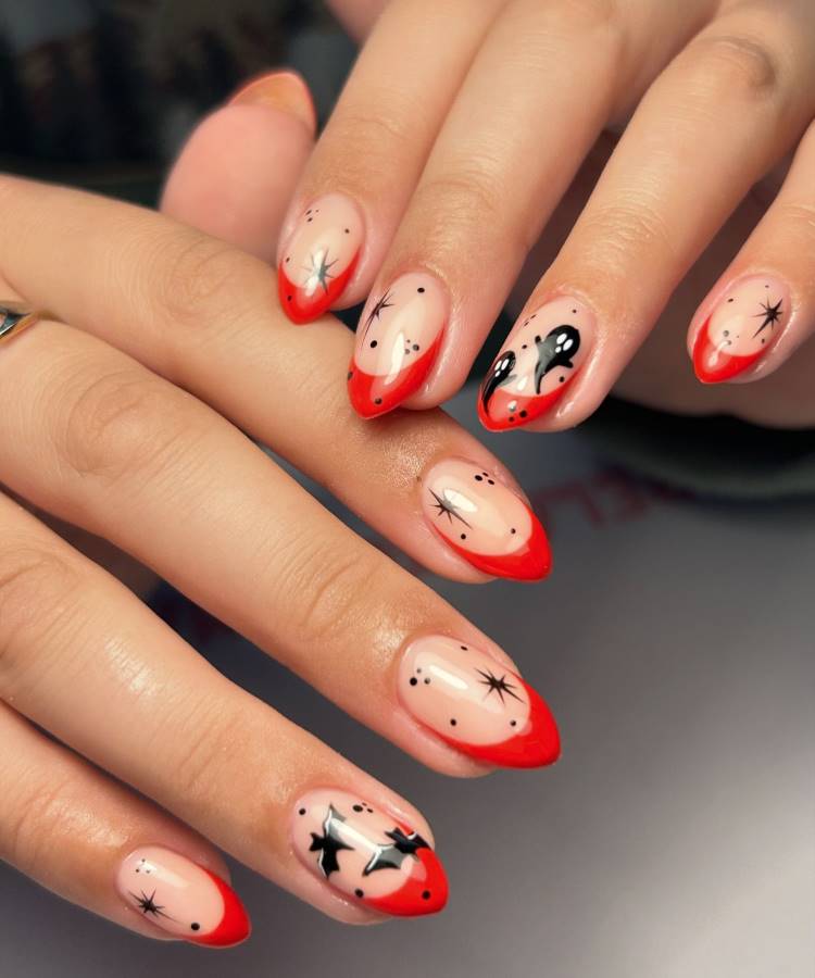 A imagem exibe unhas decoradas com pontas vermelhas e desenhos de estrelas pretas e pontos, criando um visual ousado e moderno. As unhas são de formato amendoado, com uma base clara que destaca os detalhes artísticos. O design é ideal para quem busca um estilo marcante e criativo, perfeito para ocasiões especiais ou para um toque de personalidade no dia a dia.