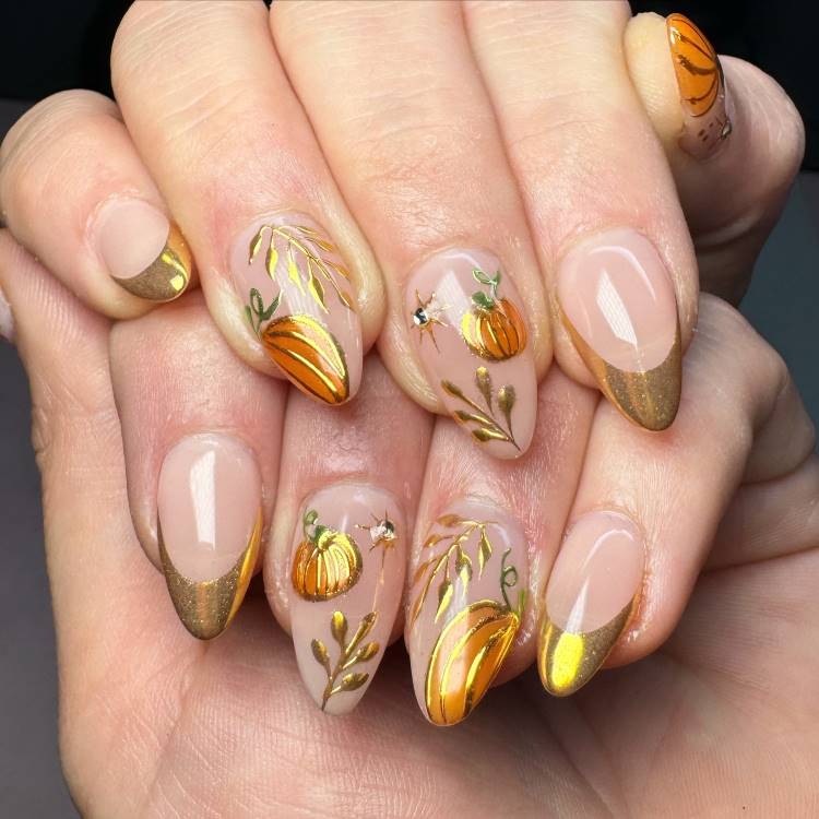 A imagem mostra unhas decoradas com um tema de outono. As unhas têm um fundo nude com pontas douradas. Desenhos de abóboras laranjas e folhas douradas adornam cada unha, criando um visual elegante e sazonal. O design é detalhado e artístico, perfeito para celebrar a temporada de colheitas e festividades de outono.