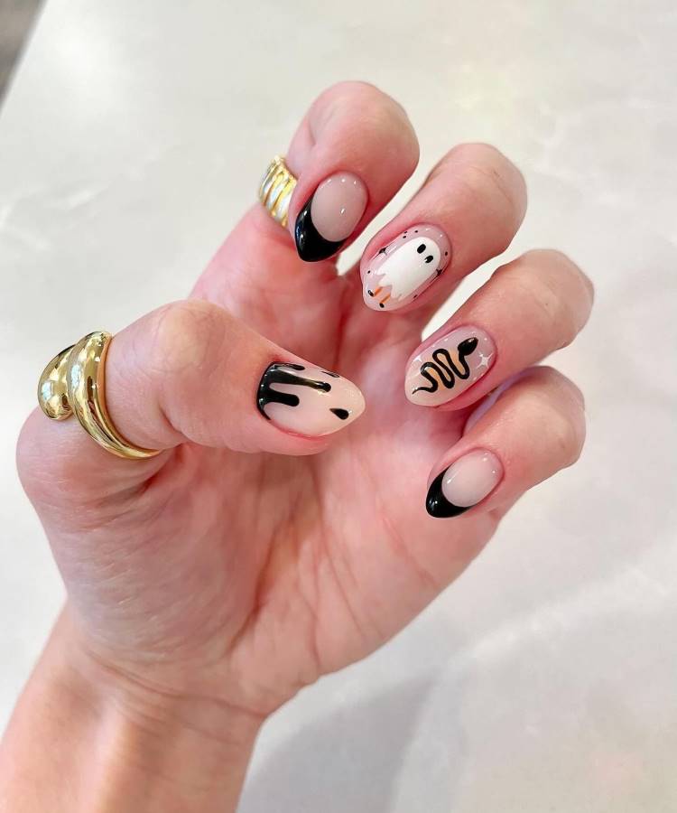 A imagem apresenta unhas decoradas com um tema de Halloween. As unhas têm pontas pretas, com detalhes como fantasmas, gotas pretas e uma cobra estilizada. O fundo é em tom nude, destacando os desenhos. O visual é criativo e festivo, ideal para a temporada de Halloween, combinando bem com acessórios dourados que complementam o estilo.