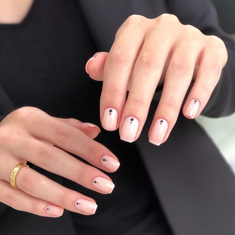 A imagem mostra mãos com unhas pintadas em um tom claro e decoradas com um pequeno ponto preto próximo à base de cada unha. Este estilo é um exemplo de unha decorada simples, que oferece um toque minimalista e elegante. As mãos estão descansando sobre um fundo escuro, destacando ainda mais a simplicidade e sofisticação do design.