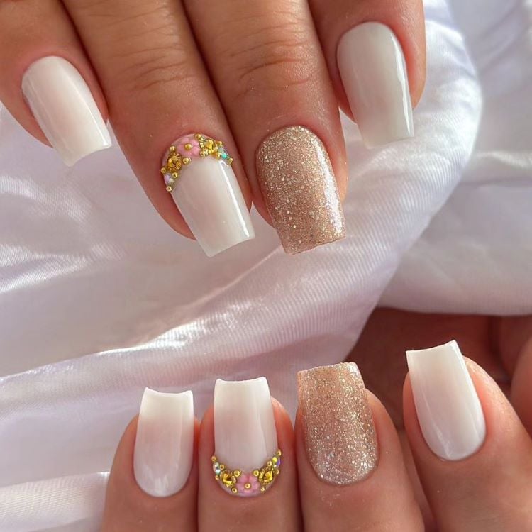 A imagem mostra uma mão com unhas decoradas de forma elegante e moderna. As unhas são pintadas em um tom claro, com uma delas coberta por glitter dourado, enquanto outra possui pequenas aplicações de flores e pedrinhas douradas perto da cutícula. Esse estilo é um exemplo de unha decorada simples, combinando sofisticação com detalhes sutis.