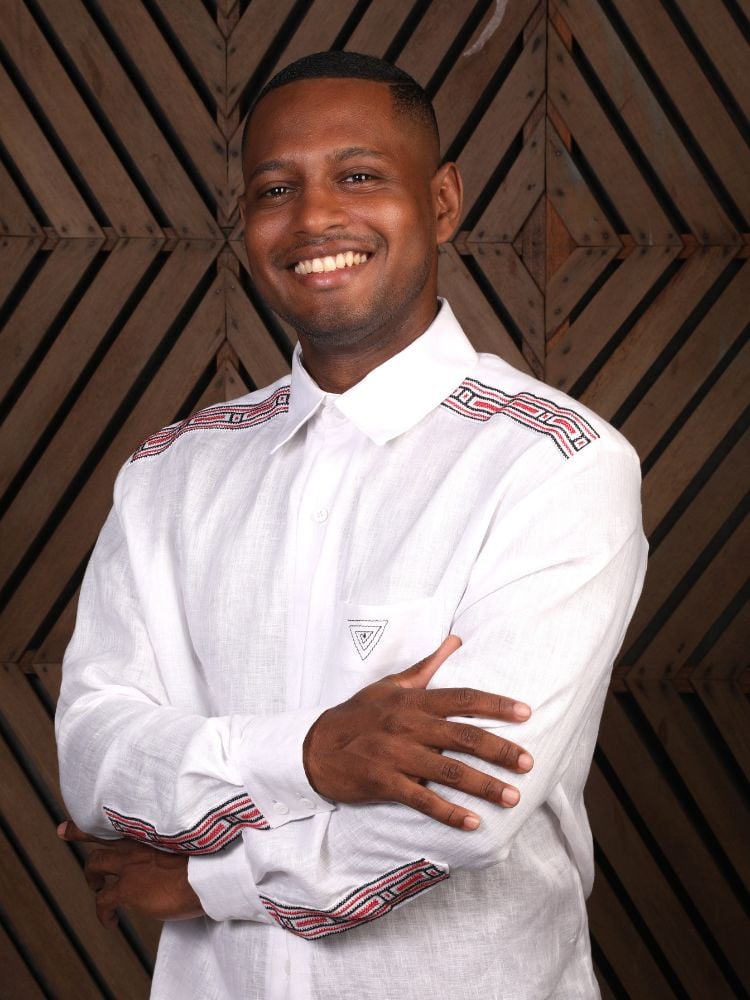 Homem de pele negra, sorrindo de braços curzados e usando camisa branca com bordados vermelhos e pretos 