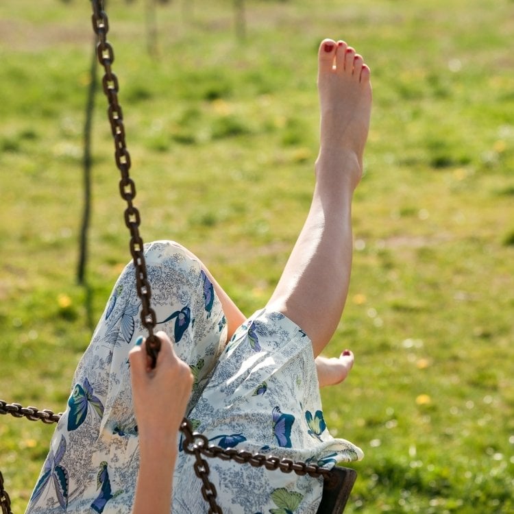 A imagem mostra uma pessoa relaxando em um balanço ao ar livre. Ela está usando um vestido com estampas florais e tem uma perna esticada, com os pés descalços. O fundo é um gramado verde, iluminado pela luz do sol.