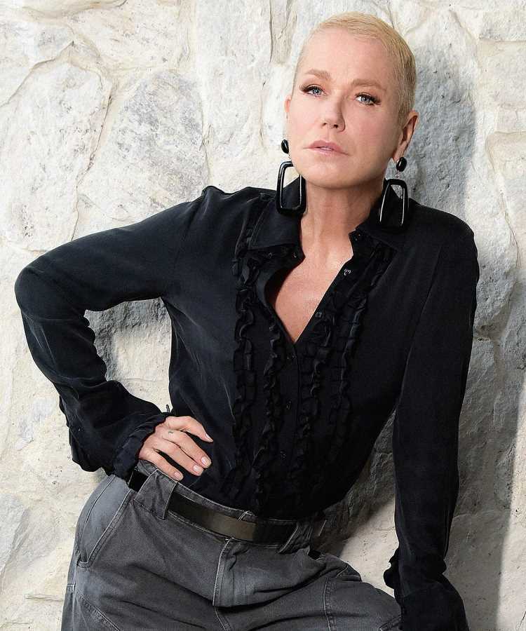 Foto de Xuxa Meneghel usando roupa preta e calça jeans com brincos grandes. Ela está com a mão na cintura encostada em uma parede de pedra clara.