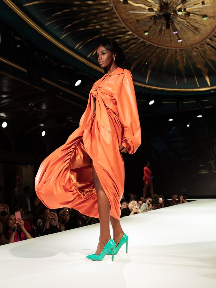 Modelo negra em passarela usando vestido mídi laranja e scarpin verde