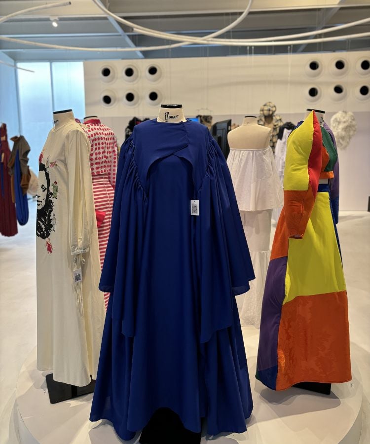 A imagem mostra uma exposição de moda brasileira com quatro manequins exibindo diferentes estilos. No centro, destaca-se um vestido azul royal volumoso. À esquerda, há uma peça branca com estampa facial e outra vermelha e branca listrada. À direita, um vestido colorido com blocos geométricos em tons vibrantes. O ambiente é moderno, com paredes brancas e iluminação embutida no teto, típico de um espaço expositivo.