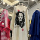 A imagem mostra três manequins exibindo diferentes estilos de vestidos em uma exposição de moda. O vestido central é branco com uma estampa facial em preto, possivelmente de Bob Marley. À esquerda, há um vestido vermelho e branco listrado com bolinhas. À direita, um vestido azul royal. O ambiente parece ser um espaço de exposição com iluminação superior.