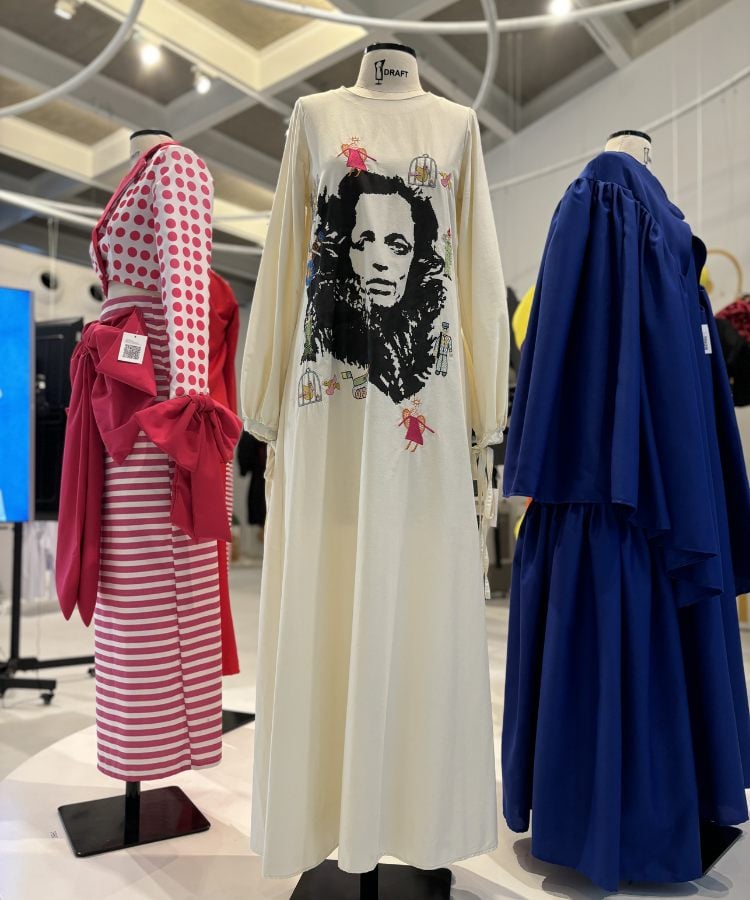 A imagem mostra três manequins exibindo diferentes estilos de vestidos em uma exposição de moda. O vestido central é branco com uma estampa facial em preto, possivelmente de Bob Marley. À esquerda, há um vestido vermelho e branco listrado com bolinhas. À direita, um vestido azul royal. O ambiente parece ser um espaço de exposição com iluminação superior.