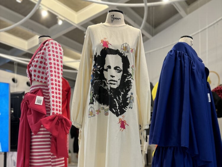 A imagem mostra três manequins exibindo diferentes estilos de vestidos em uma exposição de moda. O vestido central é branco com uma estampa facial em preto, possivelmente de Bob Marley. À esquerda, há um vestido vermelho e branco listrado com bolinhas. À direita, um vestido azul royal. O ambiente parece ser um espaço de exposição com iluminação superior.