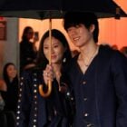 Dois modelos desfilam juntos na passarela da Amapô Jeans no SPFW N58, sob um guarda-chuva. Ambos vestem looks de jeans escuro com detalhes dourados. O ambiente é iluminado por uma luz quente, enquanto o público observa e fotografa. A cena transmite um clima de elegância e modernidade, destacando a inovação da marca.