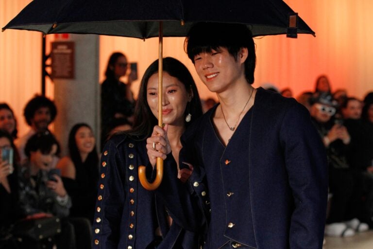 Dois modelos desfilam juntos na passarela da Amapô Jeans no SPFW N58, sob um guarda-chuva. Ambos vestem looks de jeans escuro com detalhes dourados. O ambiente é iluminado por uma luz quente, enquanto o público observa e fotografa. A cena transmite um clima de elegância e modernidade, destacando a inovação da marca.