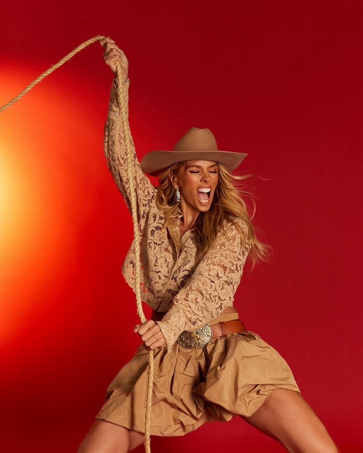 Foto de Adriane Galisteu usando chapéu de cowboy marrom, blusa de renda, saia cáqui e segurando um laço