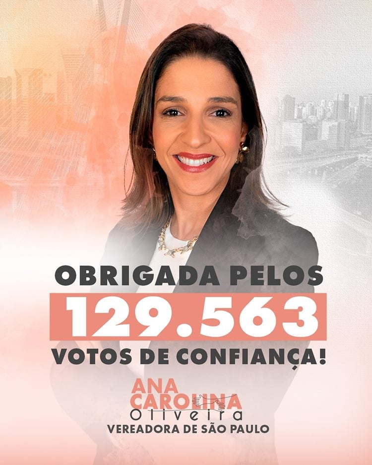 A imagem apresenta a mãe de Isabella Nardoni sorridente, com cabelo castanho e blazer preto, sobre um fundo alaranjado com a silhueta de uma cidade. Em destaque, a mensagem: "Obrigada pelos 129.563 votos de confiança!", seguida do nome "Ana Carolina Oliveira, Vereadora de São Paulo". A composição transmite gratidão e sucesso eleitoral.