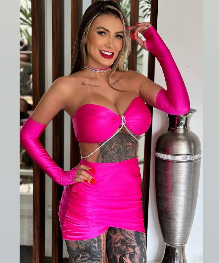 A imagem mostra uma mulher sorridente vestindo um conjunto rosa vibrante, composto por um top e uma saia curta. Ela usa luvas longas combinando e um colar brilhante. Tatuagens intricadas são visíveis em seu abdômen e pernas. Ao lado, há um vaso metálico decorativo. O fundo apresenta uma parede clara com detalhes em madeira, criando um ambiente moderno.