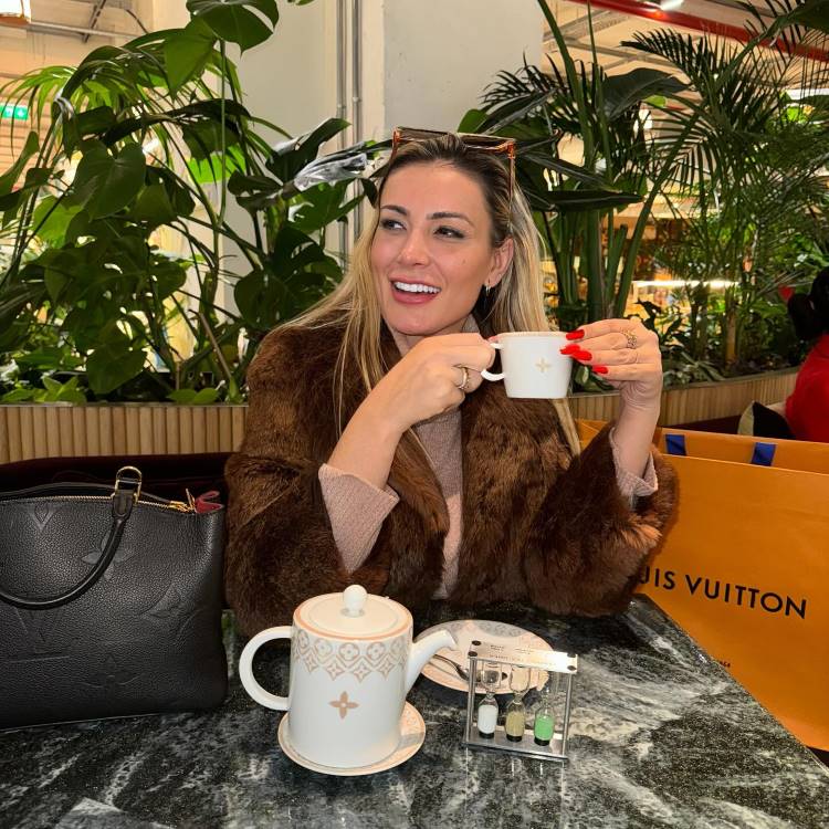 A imagem mostra uma mulher sorridente em um café elegante, segurando uma xícara de chá. Ela usa um casaco de pele marrom e tem unhas vermelhas. Ao lado, há uma bolsa preta e uma sacola da Louis Vuitton. O ambiente é decorado com plantas verdes, criando uma atmosfera sofisticada e acolhedora.