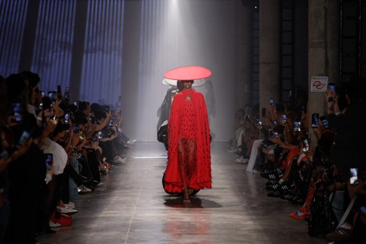 A imagem mostra uma modelo desfilando em uma passarela. Ela usa um vestido vermelho longo e transparente, decorado com detalhes escuros, e um grande chapéu vermelho de aba larga. O ambiente é escuro, com luzes focadas na passarela. Há pessoas sentadas dos dois lados, muitas segurando celulares para fotografar.