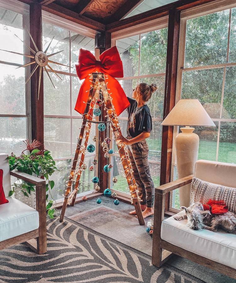 Uma mulher decora uma árvore de Natal feita com escada de madeira, iluminada por luzes e enfeitada com bolas coloridas. No topo, um laço vermelho. Ao lado, um cachorro descansa em uma poltrona. O ambiente é claro, com grandes janelas e plantas.