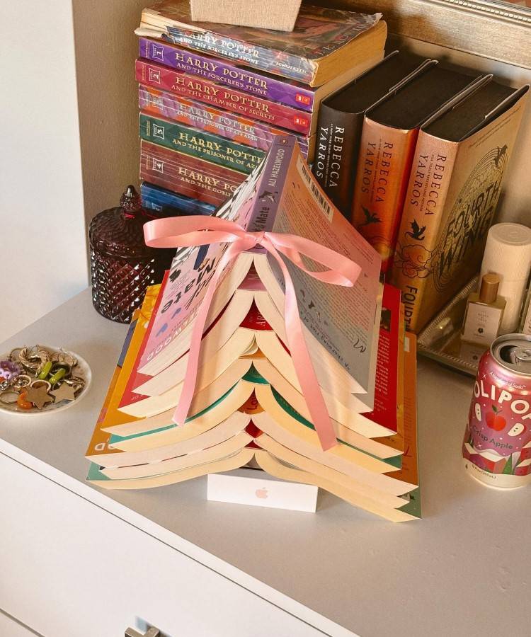 Uma árvore de Natal feita com livros abertos em formato de triângulo, decorada com uma fita rosa no topo. Ao lado, uma pilha de livros "Harry Potter" e outros volumes. Sobre a mesa, itens decorativos e uma lata de bebida.