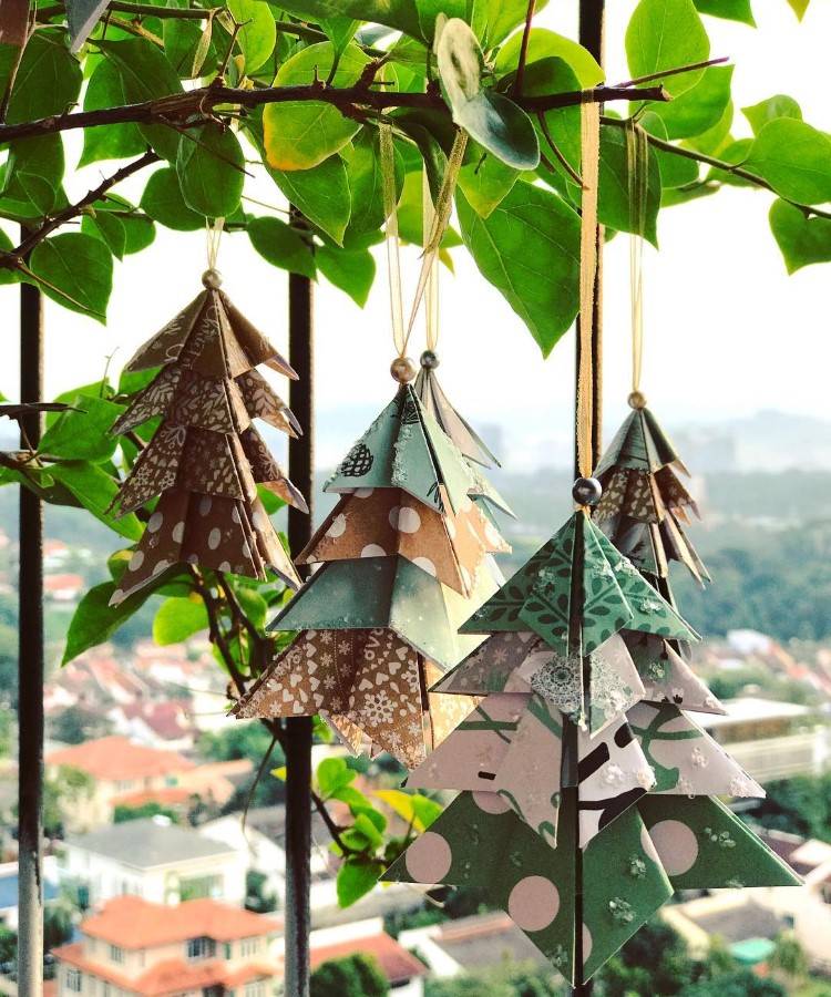 Pequenas árvores de Natal de papel penduradas em ramos de uma planta. Feitas com dobraduras de origami, exibem padrões variados em tons de verde, marrom e bege. Ao fundo, uma vista de uma cidade com telhados vermelhos e vegetação exuberante.