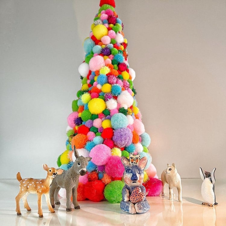 Uma árvore de Natal feita de pompons coloridos, cheia de cores vibrantes como rosa, azul, verde e amarelo. Ao redor, cinco pequenas esculturas de animais: dois veados, um coelho, um cavalo e um pinguim, criando uma cena alegre e festiva.