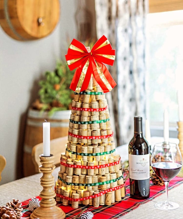 Árvore de Natal feita de rolhas de vinho, decorada com fitas vermelhas e verdes, e coroada com um grande laço vermelho. Está sobre uma mesa com toalha xadrez, ao lado de uma garrafa de vinho e uma taça cheia. Ambiente acolhedor e festivo.