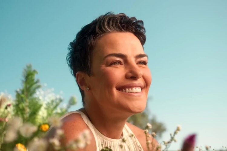 A imagem mostra uma mulher de cabelo curto e escuro (Ana Paula Arósio), sorrindo radiante em meio a um campo florido. Ela veste uma blusa clara e exibe uma expressão de serenidade sob um céu azul brilhante. Essa cena evoca a ideia de atores que desapareceram, encontrando paz e contentamento na simplicidade da natureza.