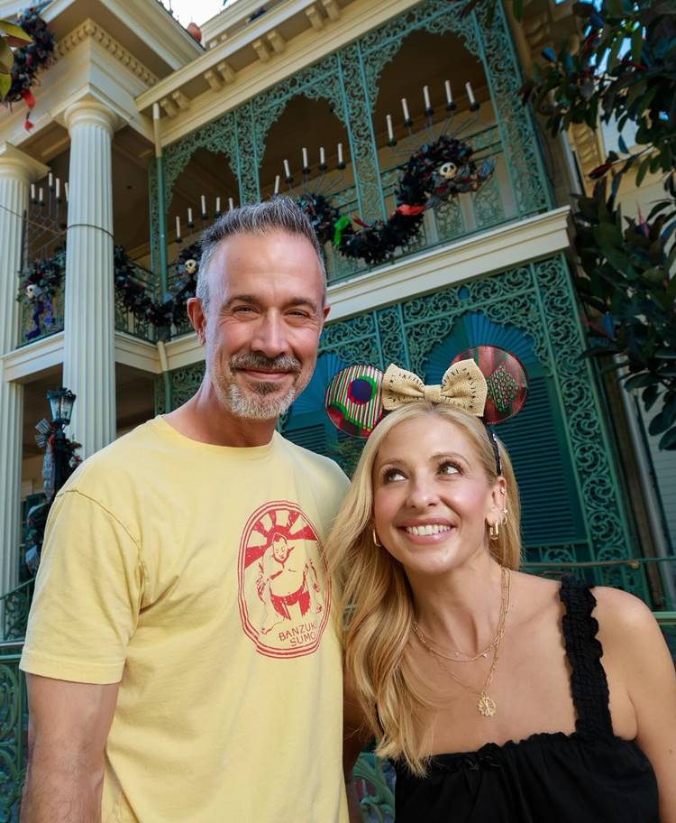 Um casal sorridente posa em frente a uma casa ornamentada com decorações festivas. O homem veste uma camiseta amarela com estampa e tem cabelo grisalho. A mulher usa uma faixa de orelhas de Minnie e um vestido preto. Ambos irradiam alegria, lembrando atores que desapareceram em algum momento, encontrando felicidade fora dos holofotes.