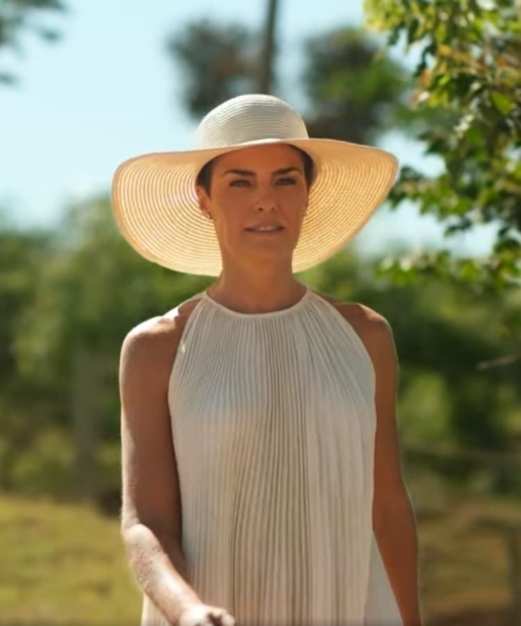 A imagem mostra uma mulher elegante (Ana Paula Arósio) em um vestido branco plissado e um chapéu de aba larga, caminhando sob a luz do sol. Ela está em um ambiente natural, com árvores ao fundo. A expressão serena sugere tranquilidade, evocando a ideia de atores que escolheram desaparecer em algum momento, encontrando paz longe dos holofotes.