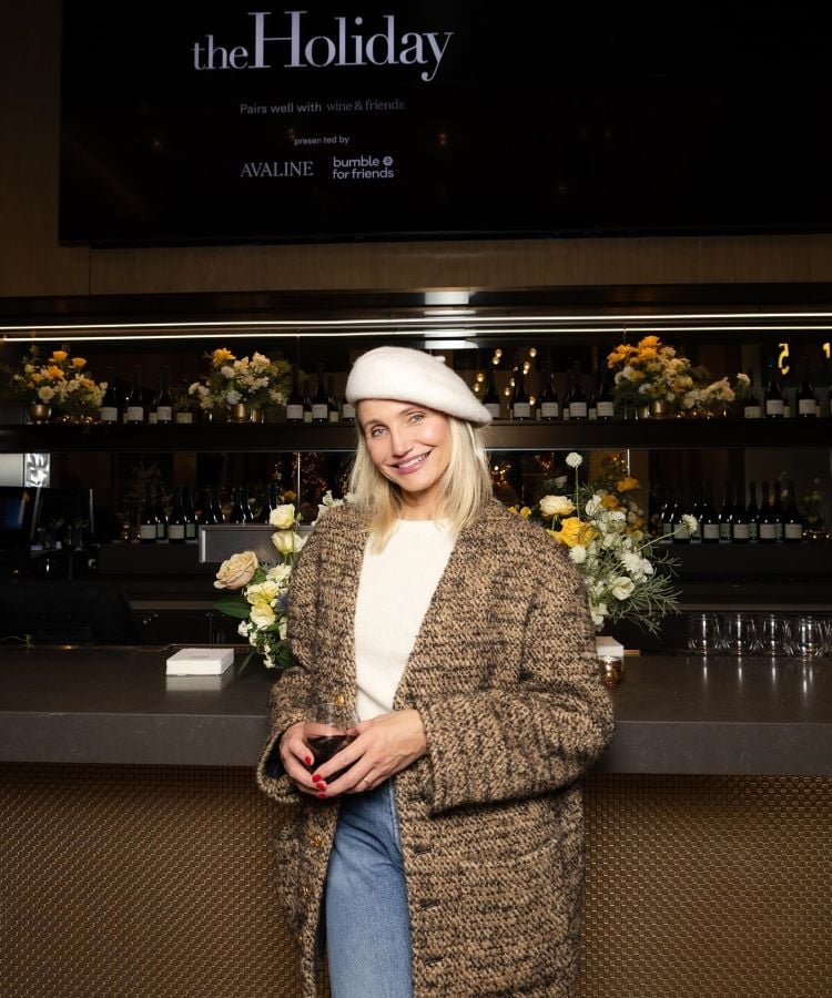 Cameron Diaz sorri em um bar elegante, vestindo um casaco marrom texturizado, blusa clara e boina branca. Ela segura uma taça de vinho tinto. Ao fundo, há arranjos de flores amarelas e brancas, com garrafas de vinho em prateleiras. Acima, uma tela exibe "the Holiday".