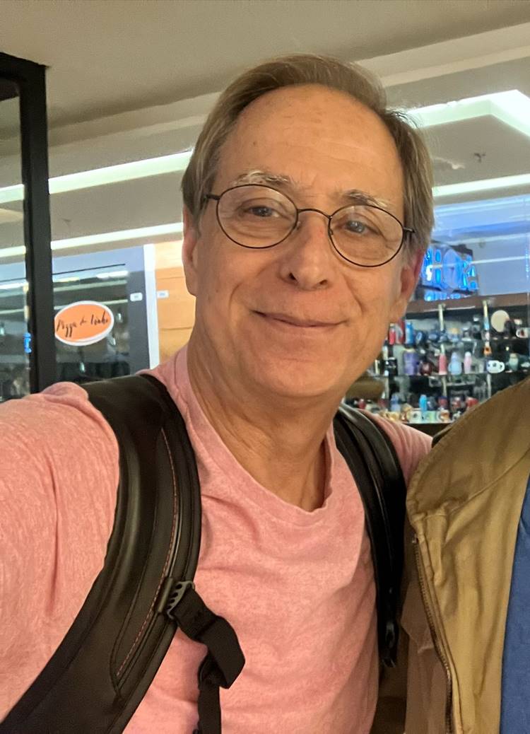 Um homem de meia-idade (Pedro Cardoso), com expressão amigável, veste uma camiseta rosa e óculos redondos. Ele carrega uma mochila preta e está em um ambiente interno, possivelmente um shopping. Seu sorriso sereno lembra aqueles atores que desapareceram em algum momento, optando por uma vida longe da fama, mas ainda radiantes de simplicidade.