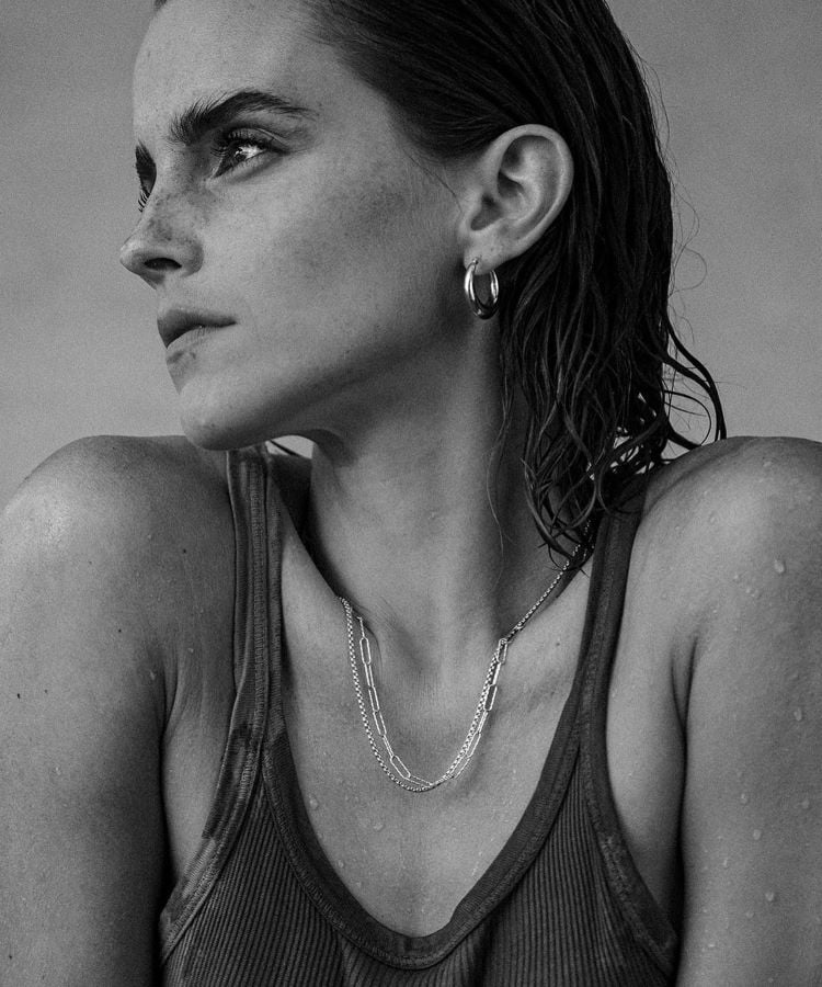 A imagem em preto e branco mostra uma mulher de perfil (Emma Watson), com cabelo molhado e penteado para trás. Ela usa uma regata justa e colares delicados. Seus olhos estão voltados para o horizonte, com uma expressão contemplativa. Ela também usa brincos de argola, e gotas de água escorrem por seu ombro.