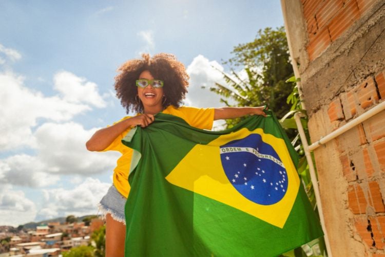 Uma mulher sorridente segura a bandeira do Brasil, destacando as cores verde, amarelo e azul com estrelas. Ela veste uma camiseta amarela e óculos de sol. Ao fundo, vê-se um céu azul com nuvens brancas e uma paisagem urbana. À direita, há uma parede de tijolos aparentes e vegetação verde. A cena transmite alegria e patriotismo.