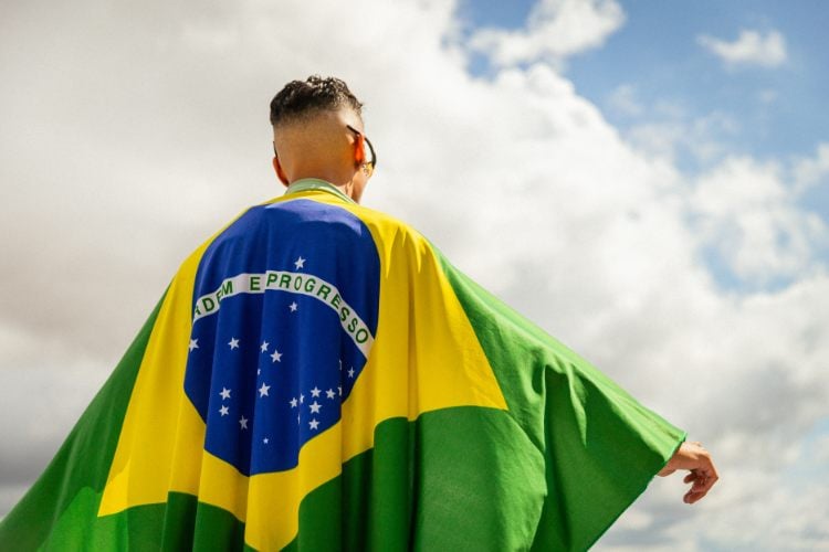 Um homem de costas veste a bandeira do Brasil como uma capa. A bandeira mostra as cores verde, amarelo, azul e branco, com o lema "Ordem e Progresso" visível. Ele está ao ar livre, sob um céu parcialmente nublado, transmitindo uma sensação de liberdade e orgulho. O homem usa óculos escuros e o clima parece ensolarado.