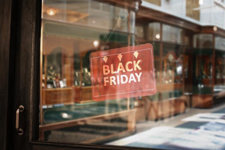 A imagem mostra uma placa vermelha pendurada em uma porta de vidro, com as palavras "Black Friday" em branco. Ao fundo, há uma loja com vitrines e prateleiras exibindo produtos. A cena reflete a típica atmosfera de promoção e expectativa de compras durante essa data especial.