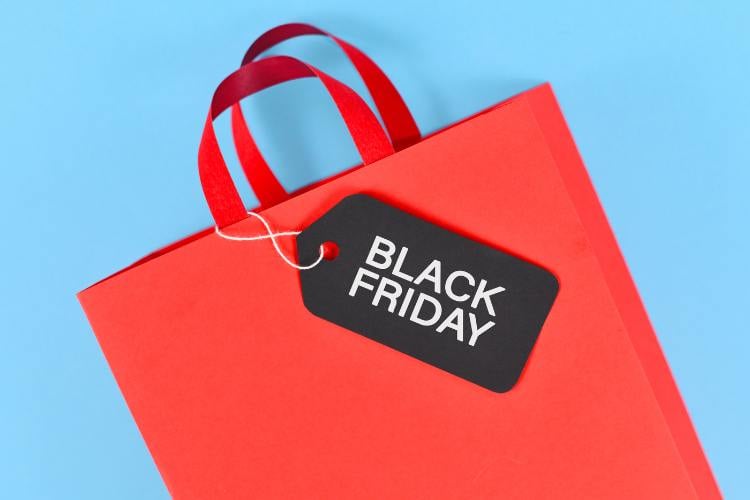 A imagem mostra uma sacola de compras vermelha com alças de fita, sobre um fundo azul claro. Preso à sacola, há um cartão preto com as palavras "Black Friday" em letras brancas. A cena simboliza promoções e compras associadas a essa data especial de vendas.