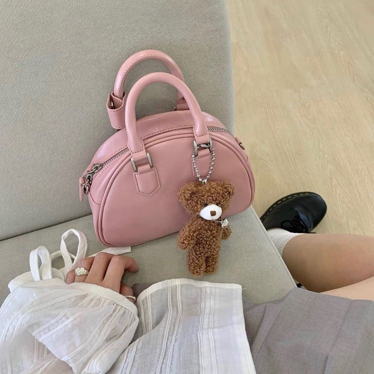 Uma bolsa rosa com alças curtas está sobre um sofá cinza. Ao lado, há um chaveiro de ursinho pendurado na bolsa. Uma pessoa, vestindo uma roupa clara e fluida, está parcialmente visível, sentada no sofá. O ambiente é iluminado e acolhedor, transmitindo um toque de delicadeza e charme.