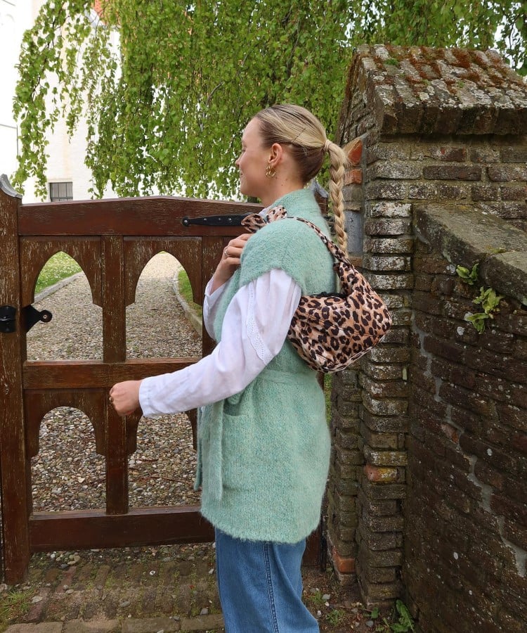 Uma mulher está diante de um portão de madeira, usando um colete verde sobre uma camisa branca. Ela carrega uma bolsa com estampa de leopardo no ombro, combinando com calças jeans. O cenário é ao ar livre, com árvores ao fundo. As bolsas com estampas animais são tendências em bolsas para 2025, destacando-se pela ousadia.