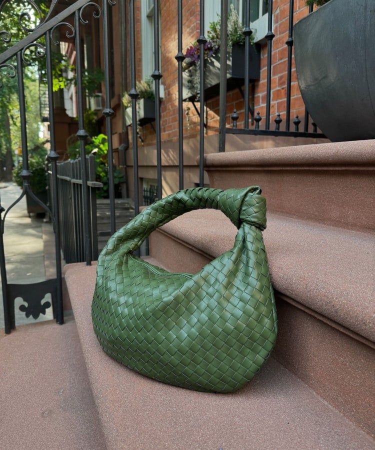 Uma bolsa verde-oliva com textura trançada está sobre degraus de pedra em um ambiente urbano. O design da bolsa é curvo e elegante, refletindo as tendências em bolsas para 2025, que valorizam texturas artesanais e tons naturais. O cenário ao ar livre, com plantas ao fundo, destaca a beleza do acessório.