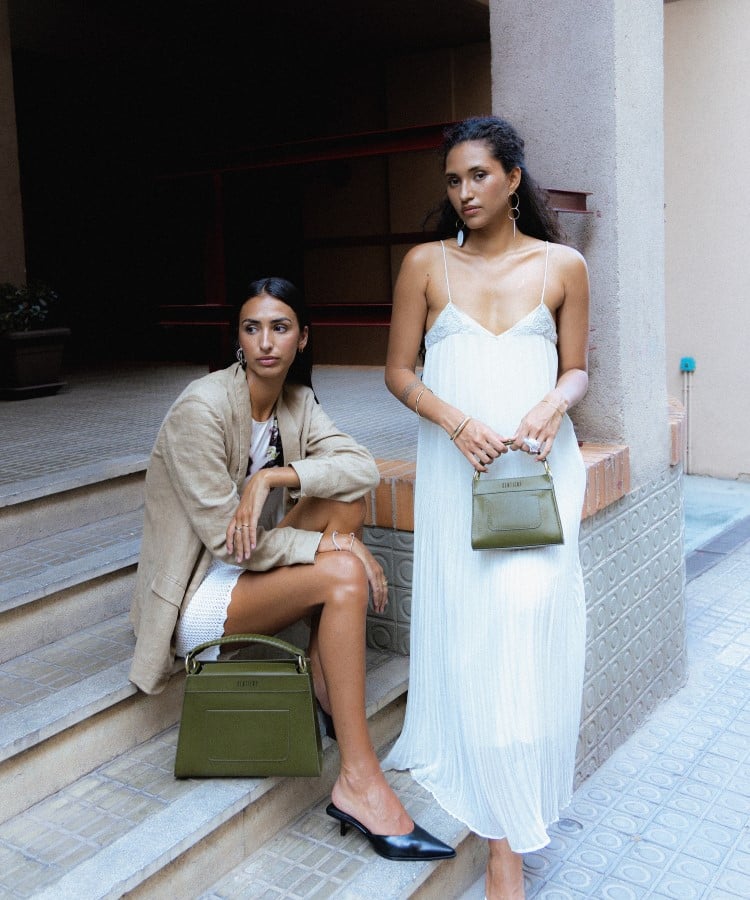 Duas mulheres posam em um ambiente urbano. Uma veste um casaco bege e carrega uma bolsa verde-oliva estruturada. A outra usa um vestido branco fluido e segura uma bolsa menor no mesmo tom. As tendências em bolsas para 2025 incluem cores naturais e designs estruturados, conferindo sofisticação e modernidade ao visual.