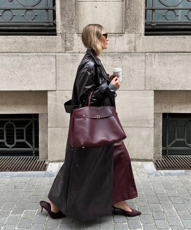 Uma mulher caminha em uma rua urbana, vestindo um longo casaco de couro preto e uma saia marrom. Ela segura uma bolsa grande e extravagante em tom bordô, que complementa seu look sofisticado. As tendências em bolsas para 2025 destacam peças de tamanho exagerado e cores ricas, que adicionam um toque de ousadia e estilo.