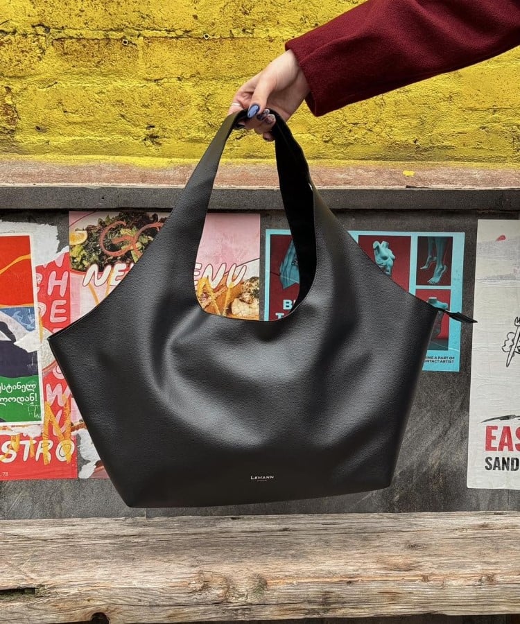 Uma bolsa preta de couro com design curvo e alças largas é exibida em frente a um mural colorido. O fundo amarelo vibrante contrasta com a simplicidade elegante da bolsa. As tendências em bolsas para 2025 incluem formas esculturais e minimalistas, que se destacam em qualquer ambiente urbano, trazendo modernidade e funcionalidade.