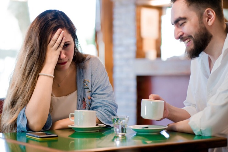 Mulher de pele clara usando blusa branca e jaqueta azul com expressão entediada e mão no rosto, sentada à mesa com homem de pele clara que parece feliz