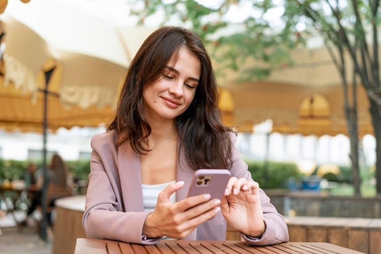 Mulher de pele clara usando blazer lilás e blusa branca, sentada à mesa de madeira, mexendo no celular com expressão feliz 