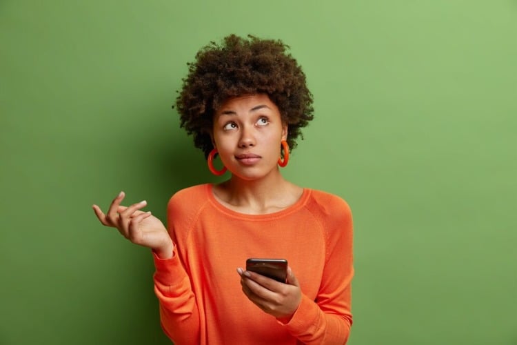 Mulher de pele negra em fundo roxo usando blusa laranja e segurando celular com expressão confusa 