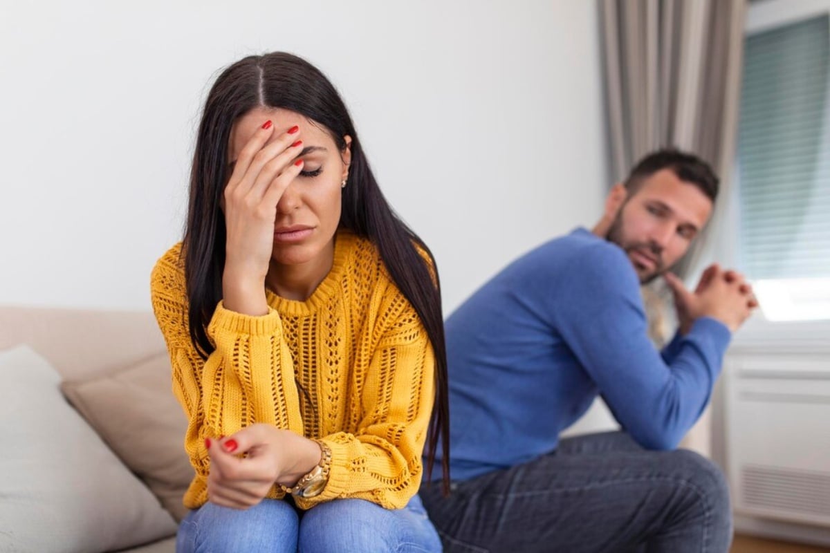 Mulher de pele clara sentada em sofá usando blusa amarela e calça jeans, com expressão triste e confusa, de costas para homem de pele clara usando blusa azul e calça cinza com expressão triste