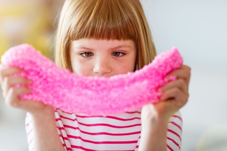 A imagem mostra uma criança concentrada segurando uma massa sensorial rosa, esticando-a com as mãos. Esse tipo de massa é um dos presente para criança com autismo, pois ajuda a desenvolver habilidades motoras e sensoriais. A criança está vestida com uma camiseta listrada, e o ambiente parece ser iluminado e acolhedor, propício para brincadeiras.