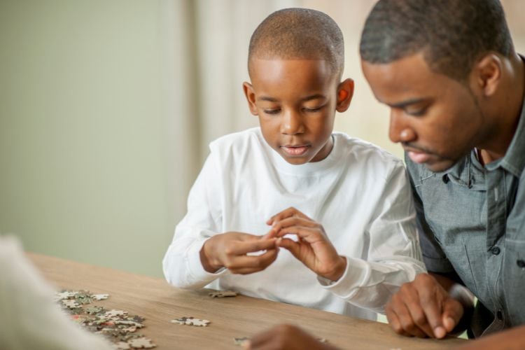 A imagem mostra um menino e um adulto montando um quebra-cabeça em uma mesa. O menino, concentrado, segura uma peça enquanto o adulto observa atentamente. Quebra-cabeças são excelentes presente para criança com autismo, ajudando no desenvolvimento cognitivo e na coordenação motora. O ambiente é calmo e propício para atividades educativas e interativas.